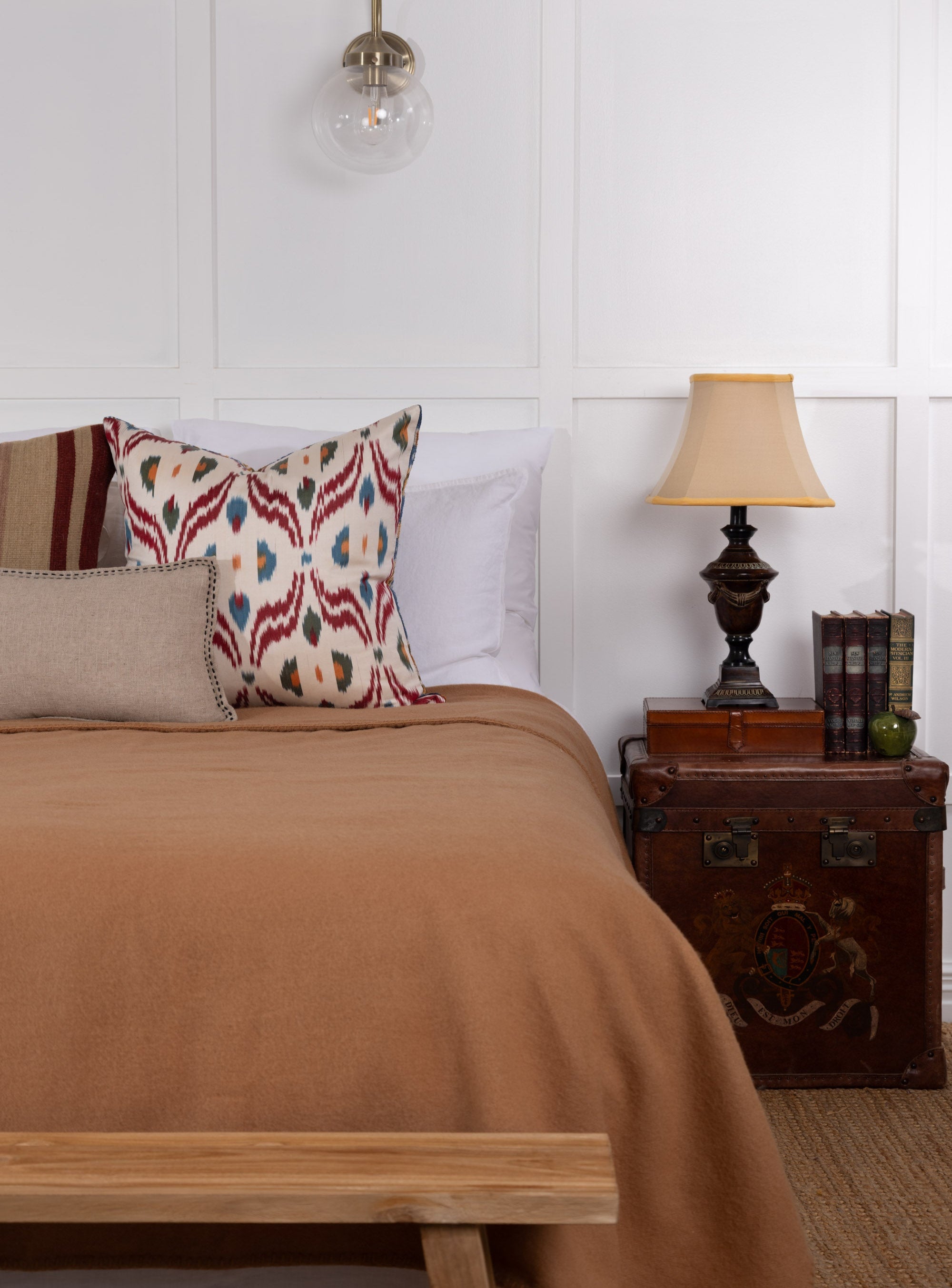 A beige Camel NZ Wool Blanket laid out on a bed in a bedroom setting, with a decorative pillow, a lamp, and a wooden side table.
