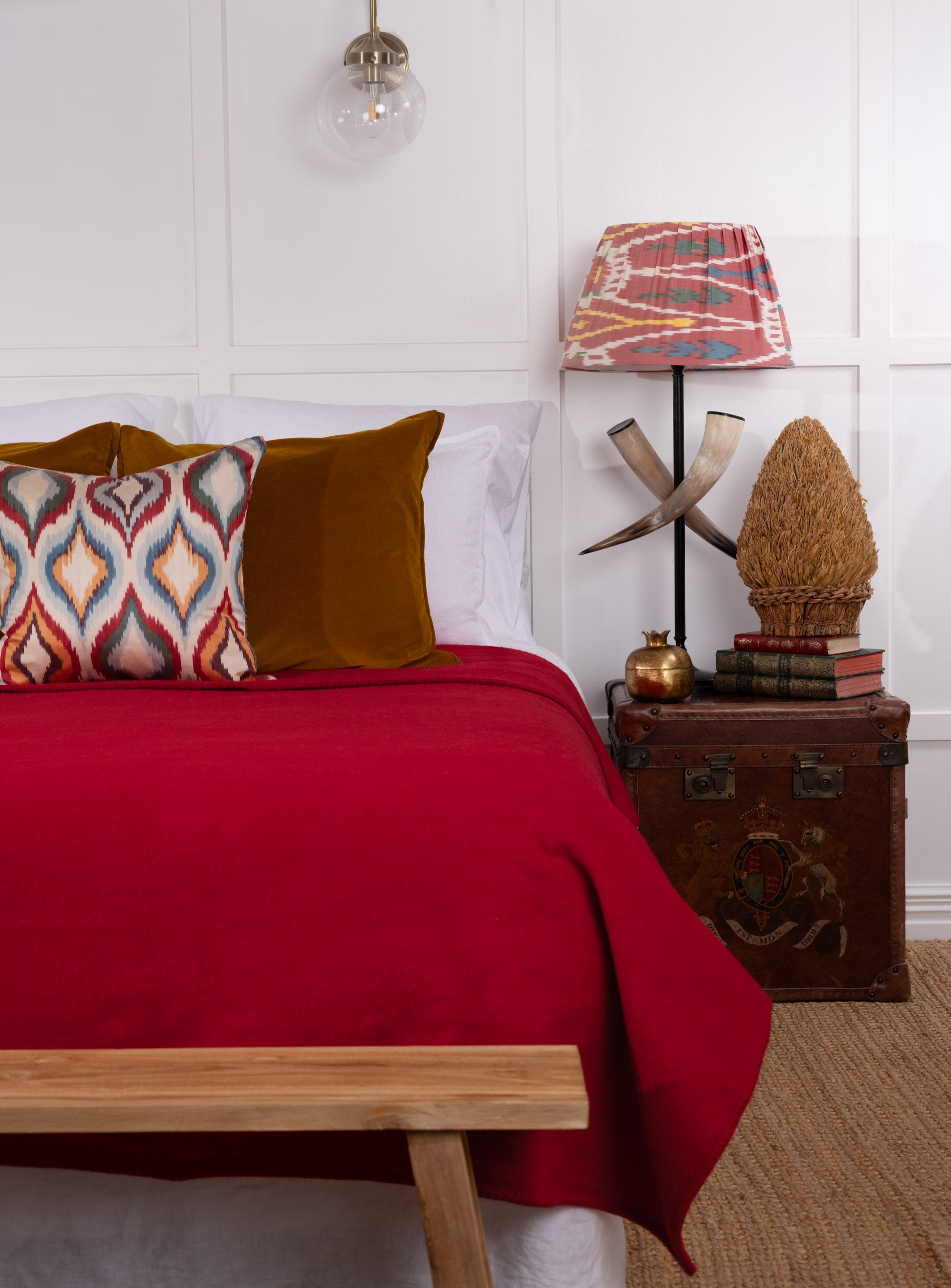 A cherry red wool blanket draped over the end of a bed with a wooden bed frame, in a room with a side table, lamp, and decorative items.