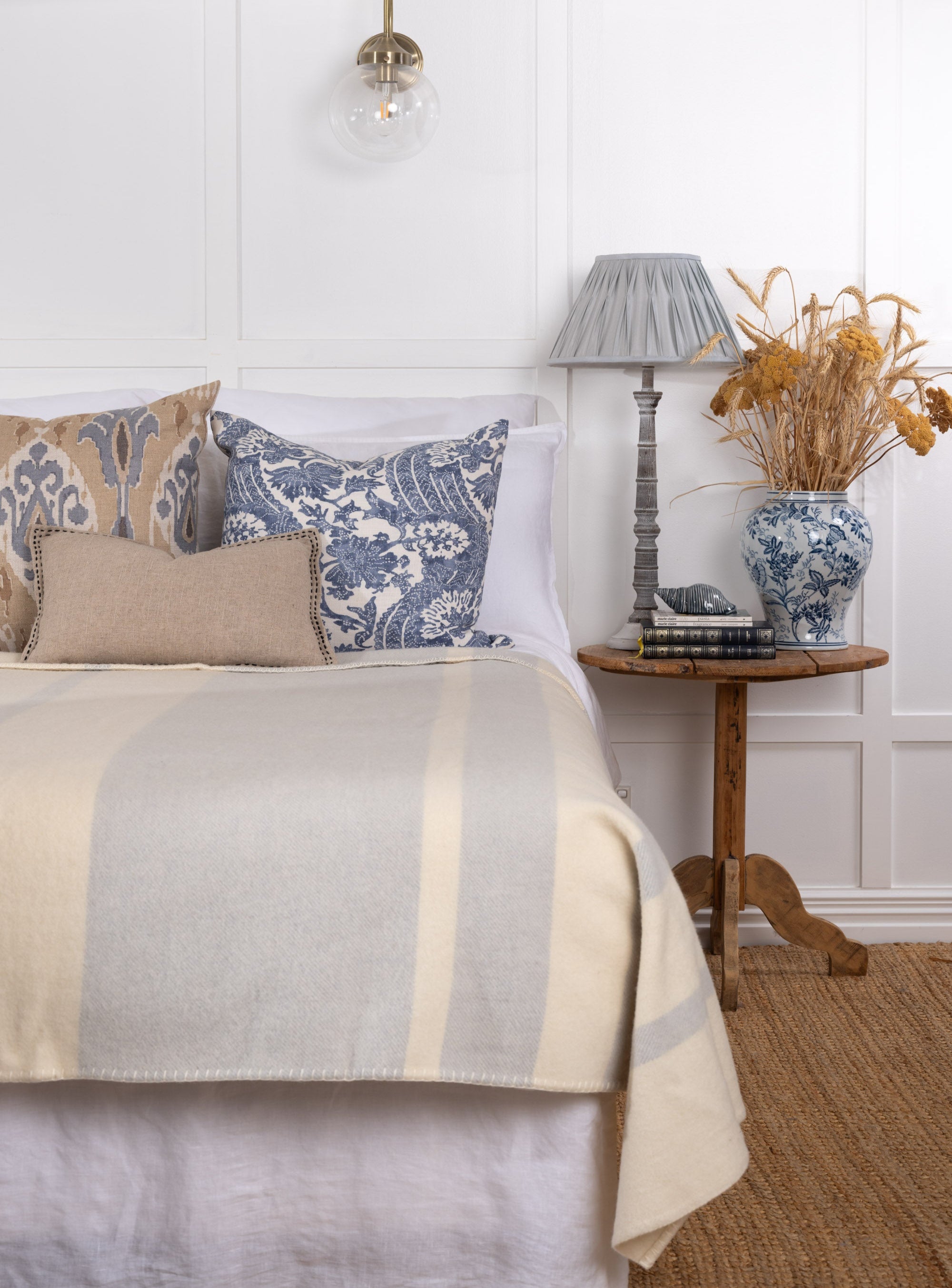 A lambswool blanket with a subtle striped pattern in white and blue shades, displayed at the foot of a bed in a styled bedroom setting.