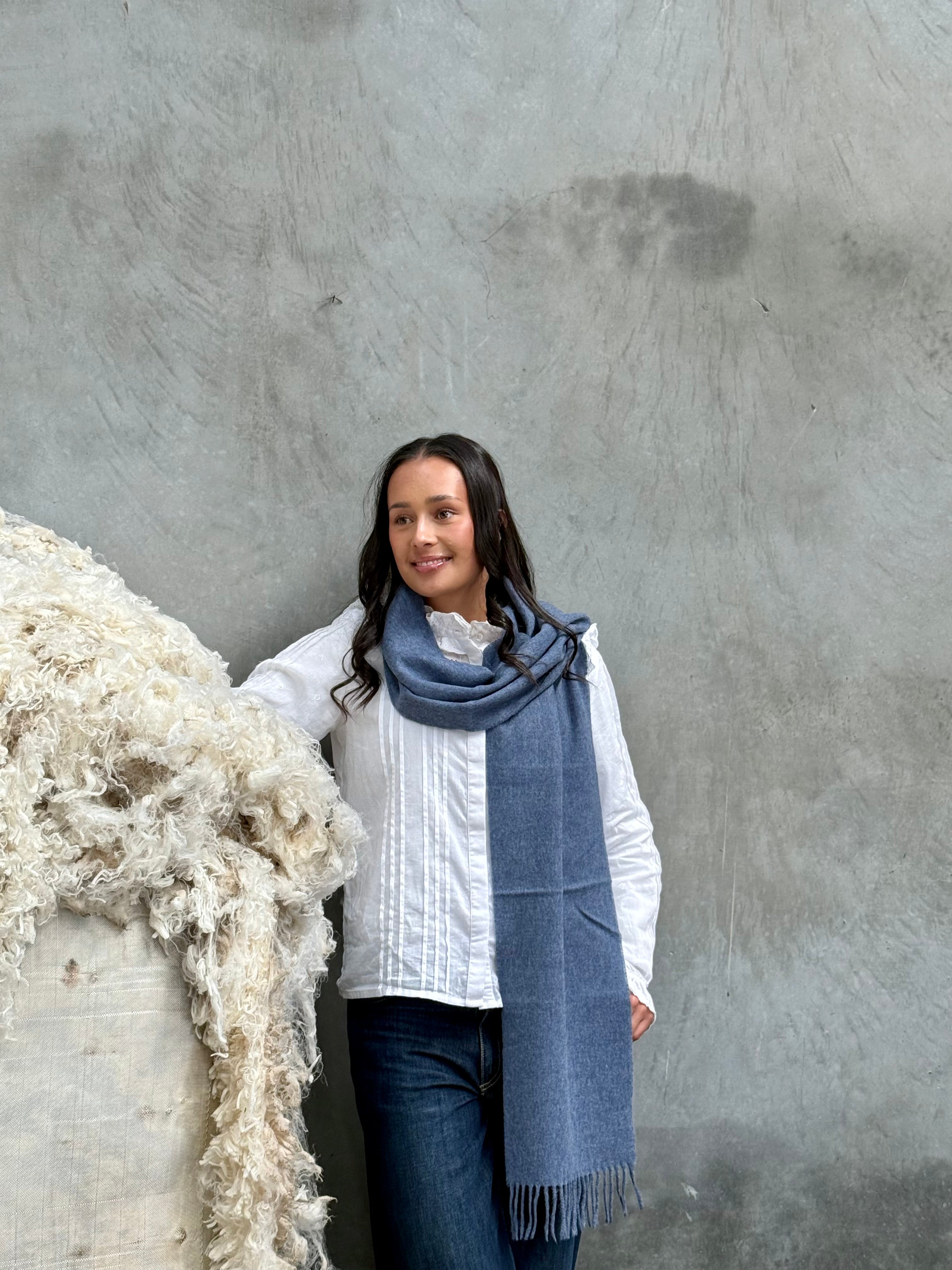 A person wearing an airforce blue merino scarf with a classic twill weave, standing next to a pile of white wool.