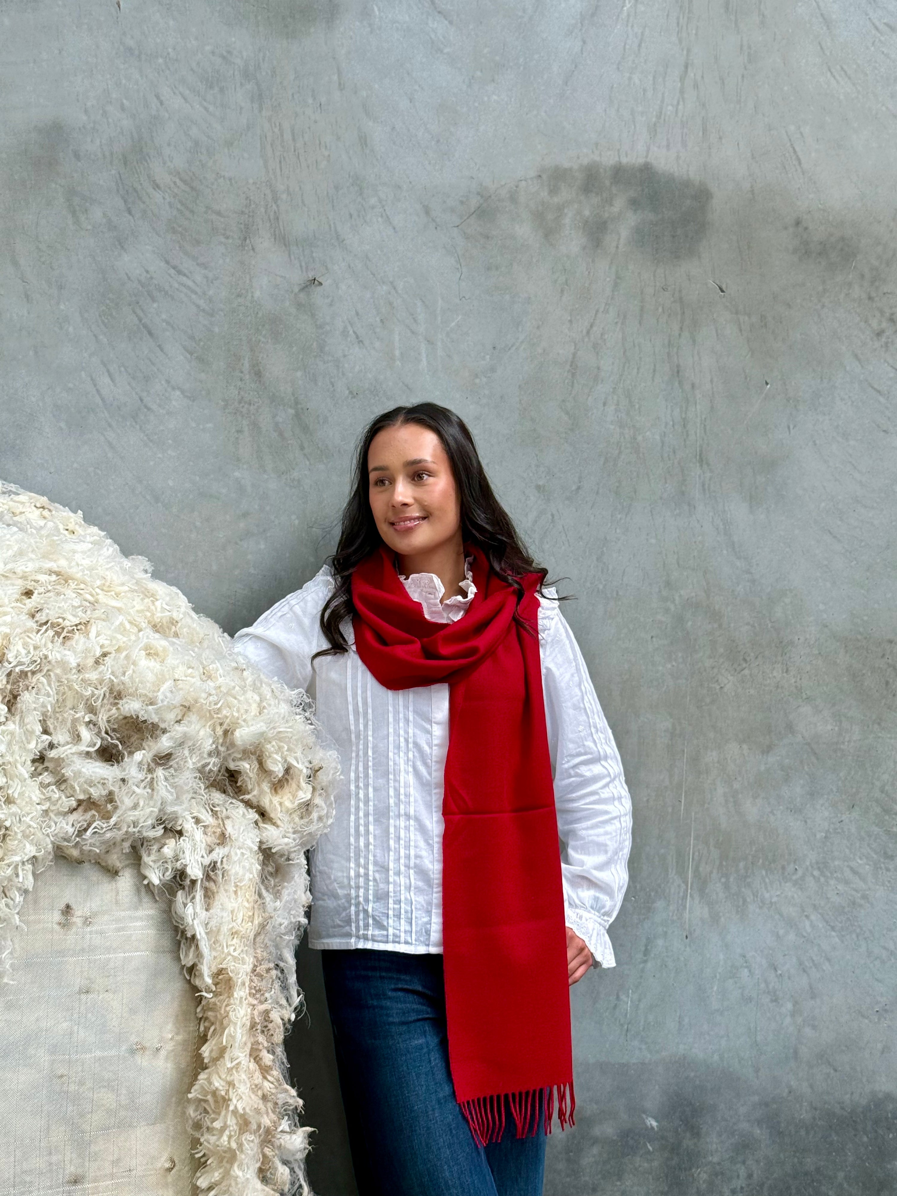 A person wearing a white shirt and jeans, accessorized with a vibrant red merino wool scarf, standing against a grey background.
