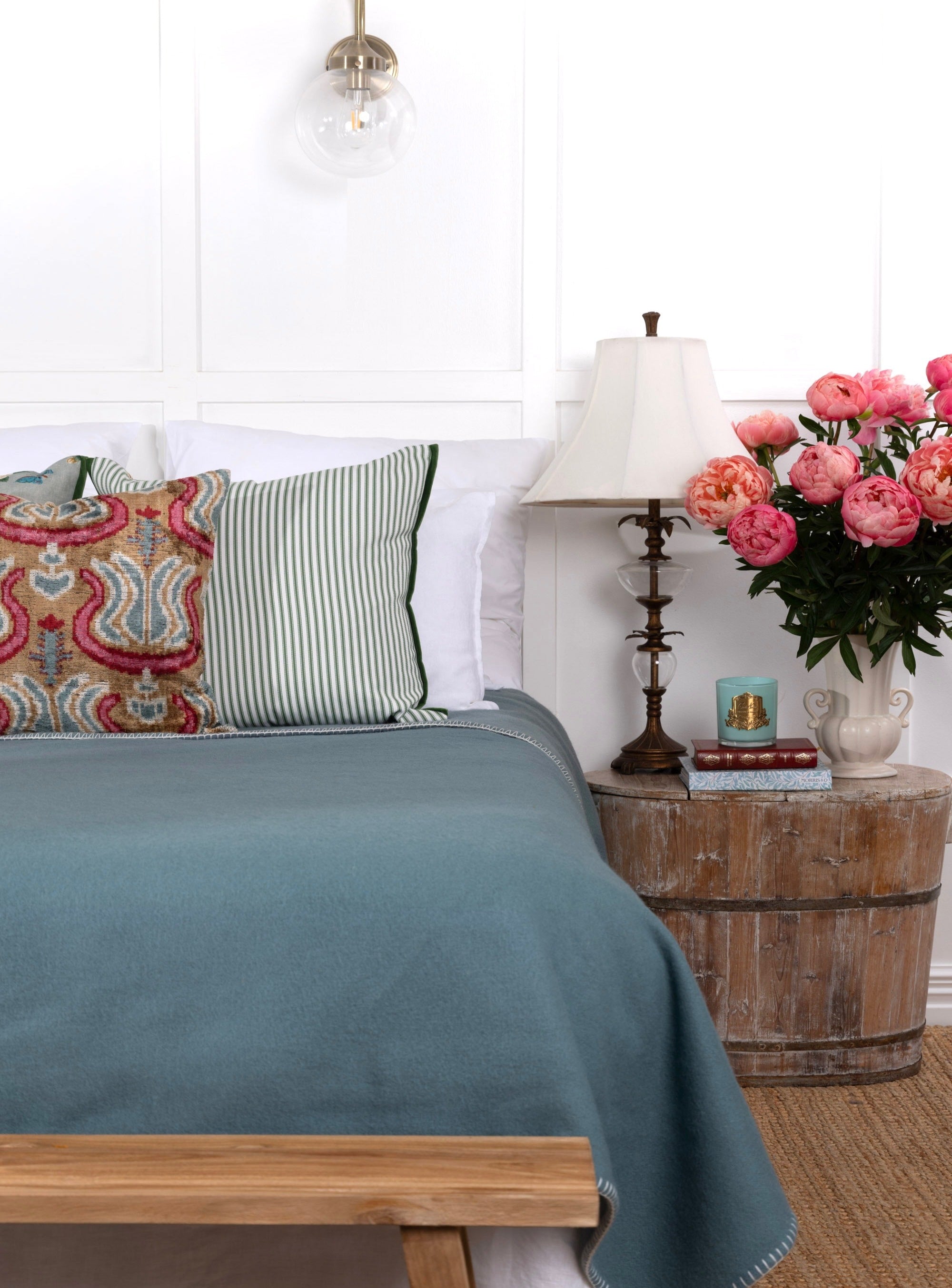 A pale sea green wool blanket laid out at the foot of a bed with decorative pillows, a lamp, and a vase of flowers in the background.