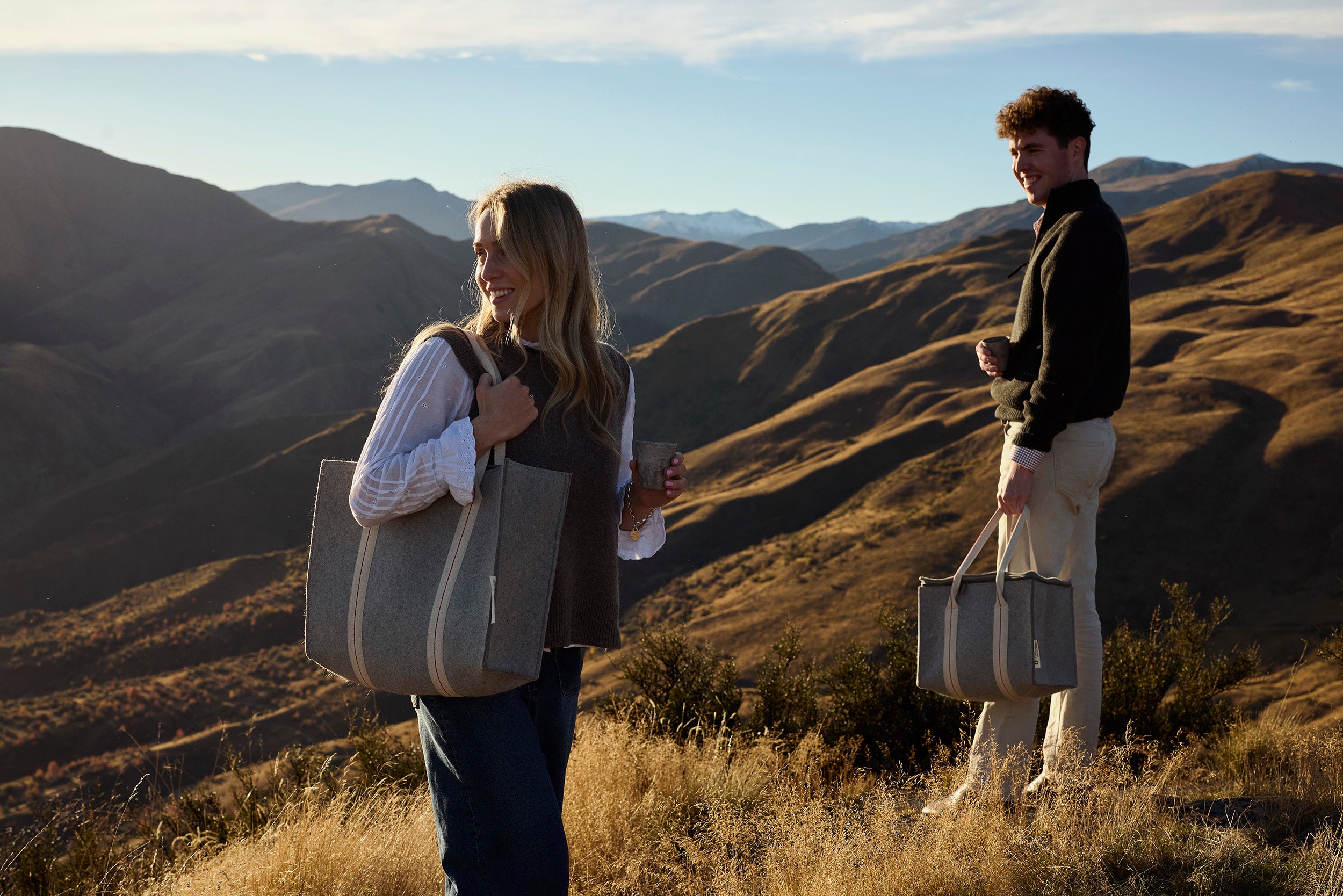 Two people standing on a grassy mountain ridge holding large gray tote bags, with rolling hills and distant mountains in the background under a blue sky.