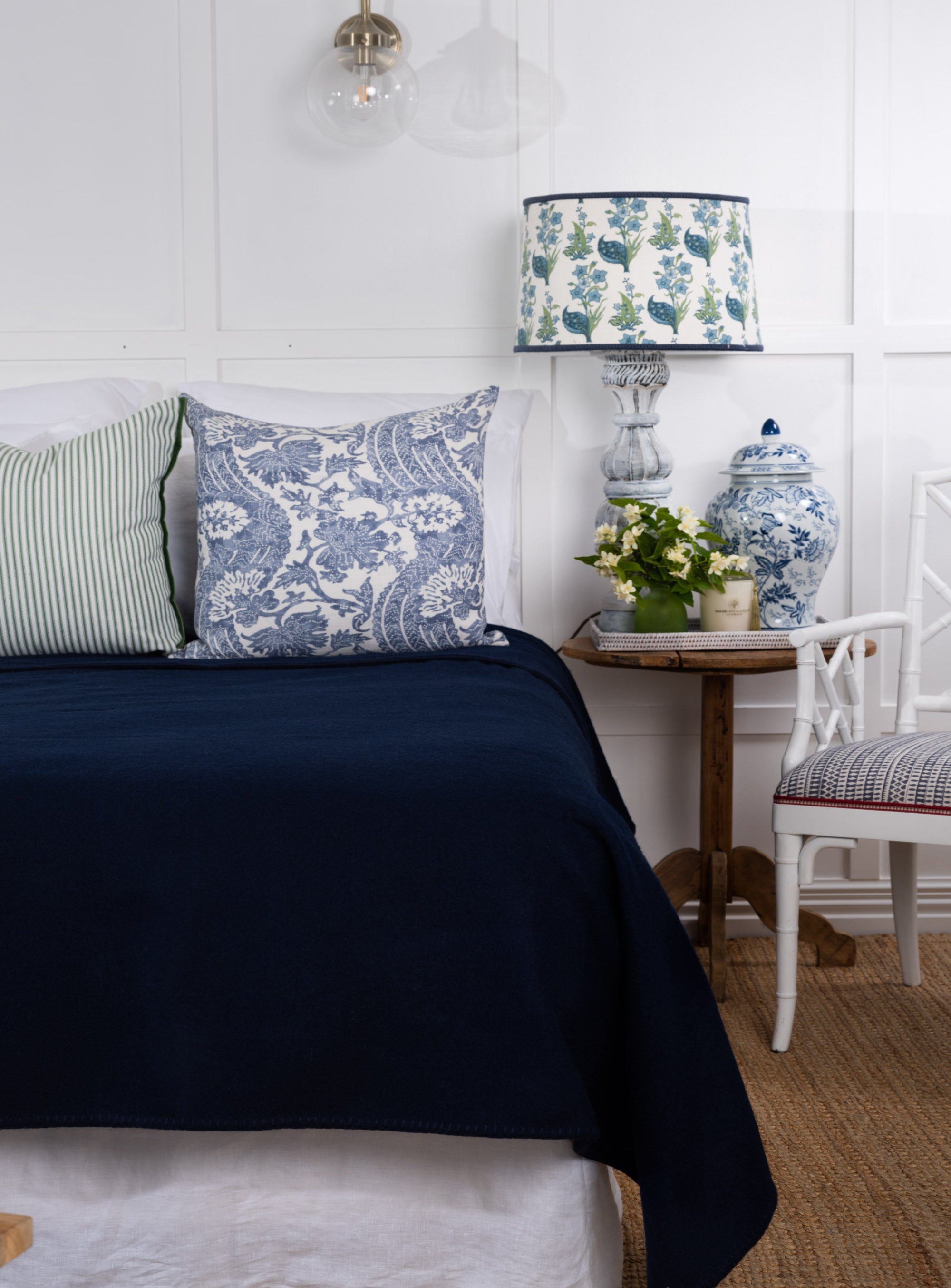 A navy New Zealand wool blanket draped over the end of a bed with a decorative pillow, in a room with a white wall and wooden flooring.
