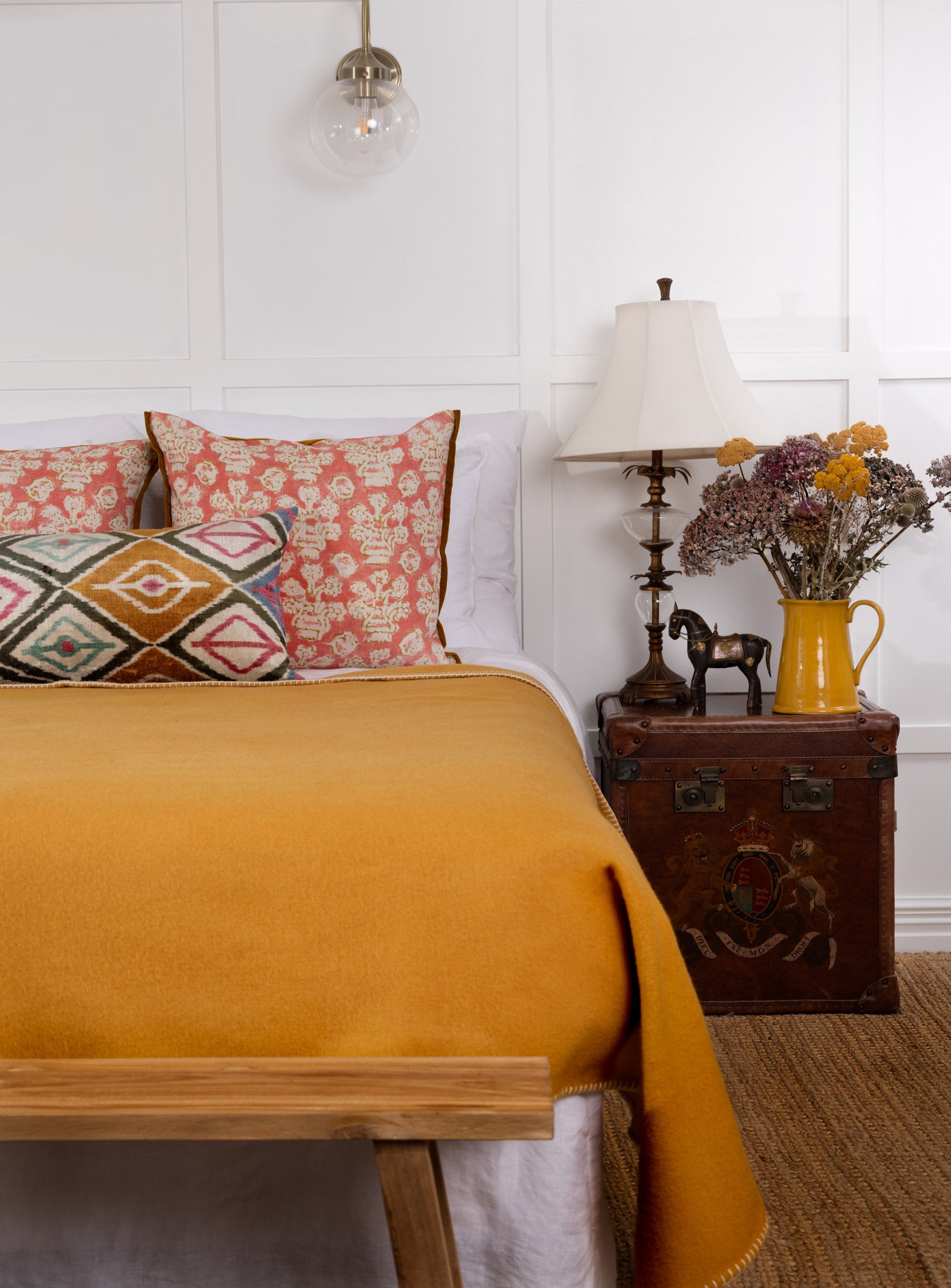 An ochre yellow wool blanket draped over the end of a bed in a room with a wooden floor and white walls, alongside decorative pillows, a lamp, and a vase with flowers.