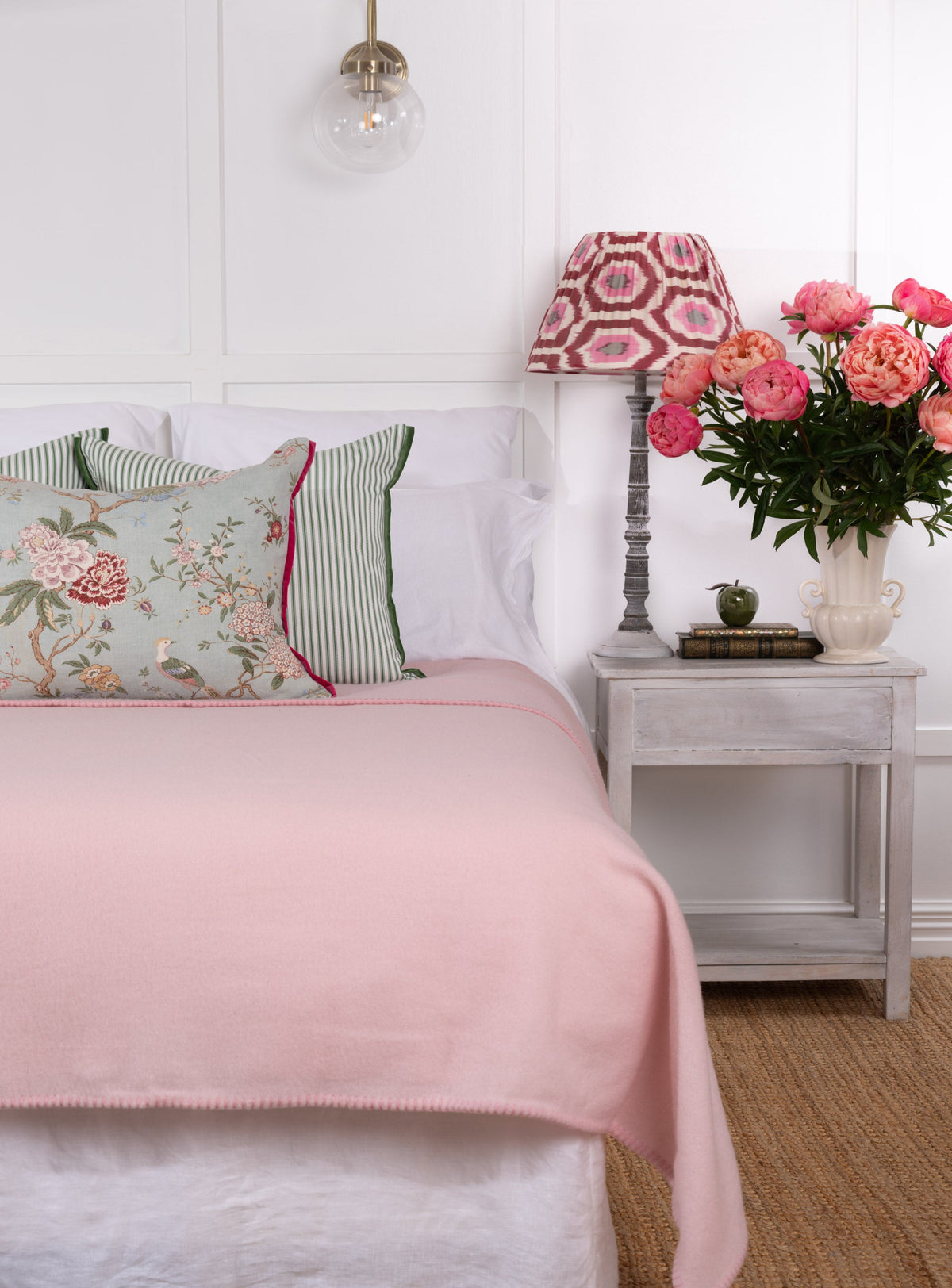 A pale pink NZ wool blanket draped over the edge of a bed, with a white and green floral pillow and a wooden side table with a lamp and a vase of pink flowers in the background.