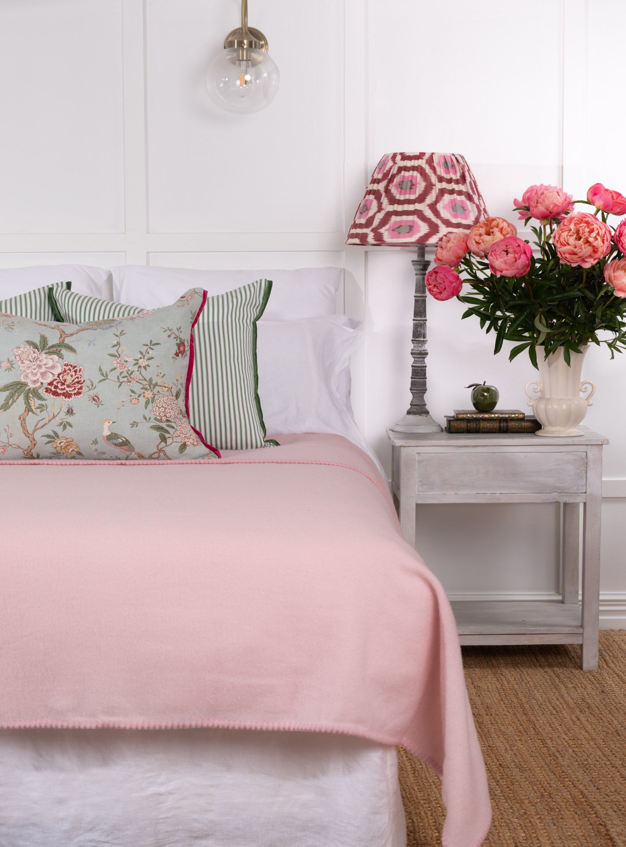 A pale pink NZ wool blanket draped over the edge of a bed, with a white and green floral pillow and a wooden side table with a lamp and a vase of pink flowers in the background.