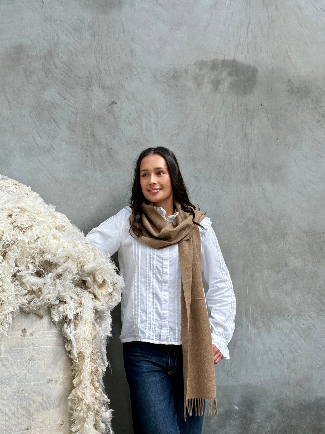 A person standing next to a large pile of wool, wearing a light brown merino scarf with a classic twill weave.