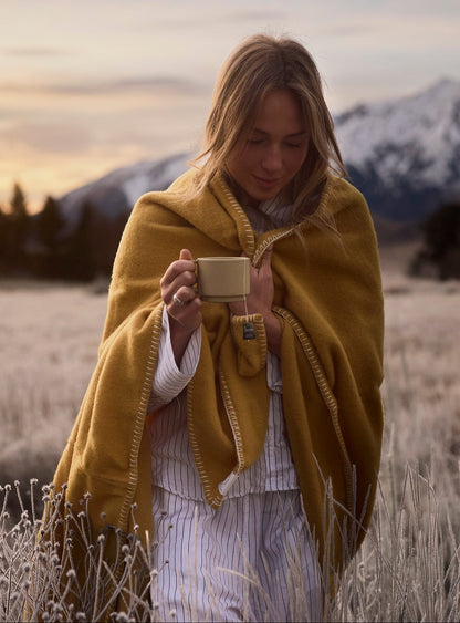 A woman wrapped in a mustard yellow blanket stands in a frosty field at sunset, holding a small beige box. Snow-capped mountains are visible in the background.