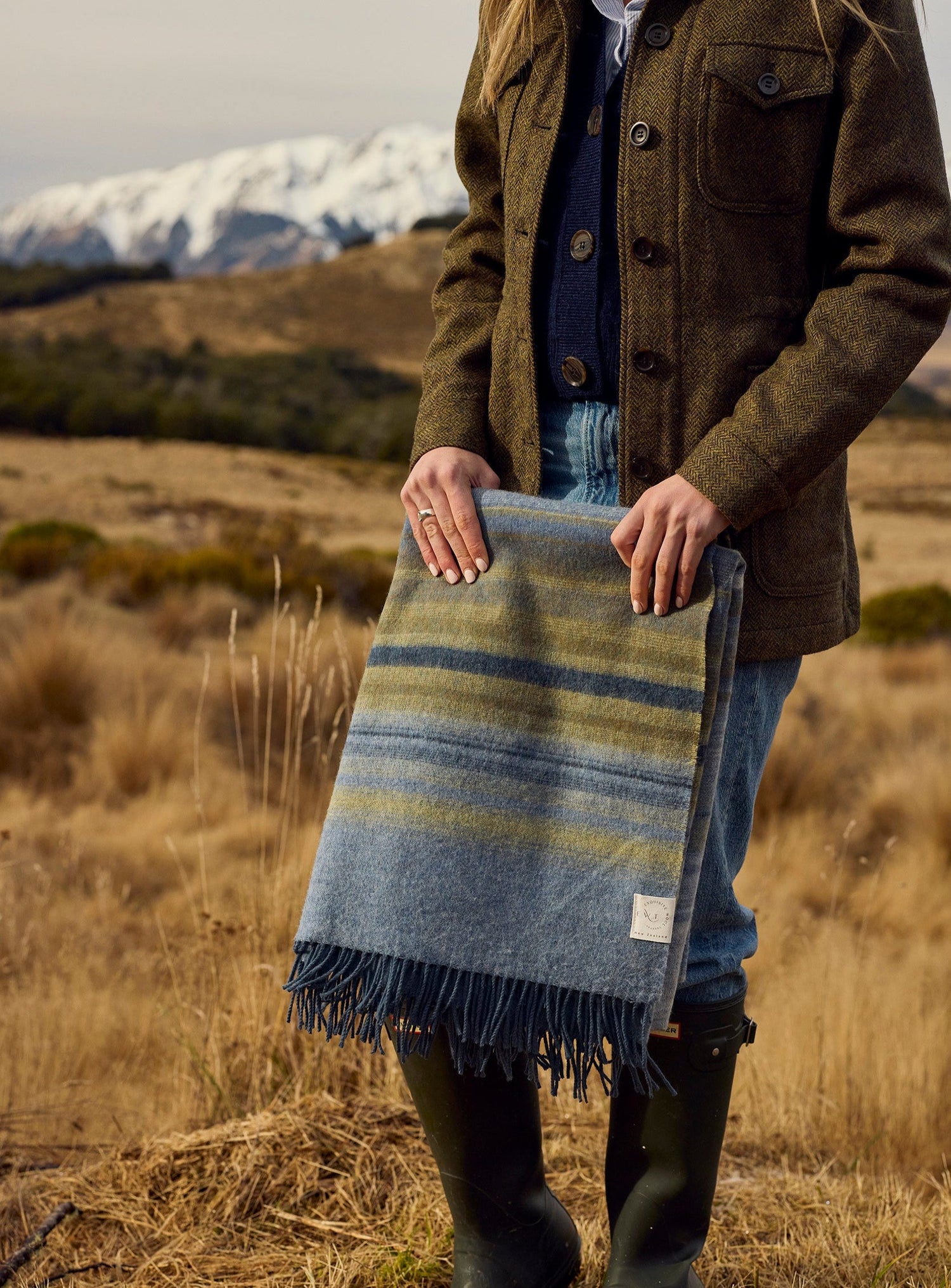 A person standing outdoors in a dry grassy field with mountains in the background, holding a folded striped wool blanket or throw in blue, gray, olive, and tan tones with dark fringe.
